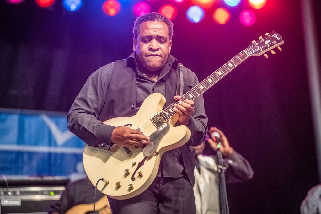 A man wearing black holding tan guitar