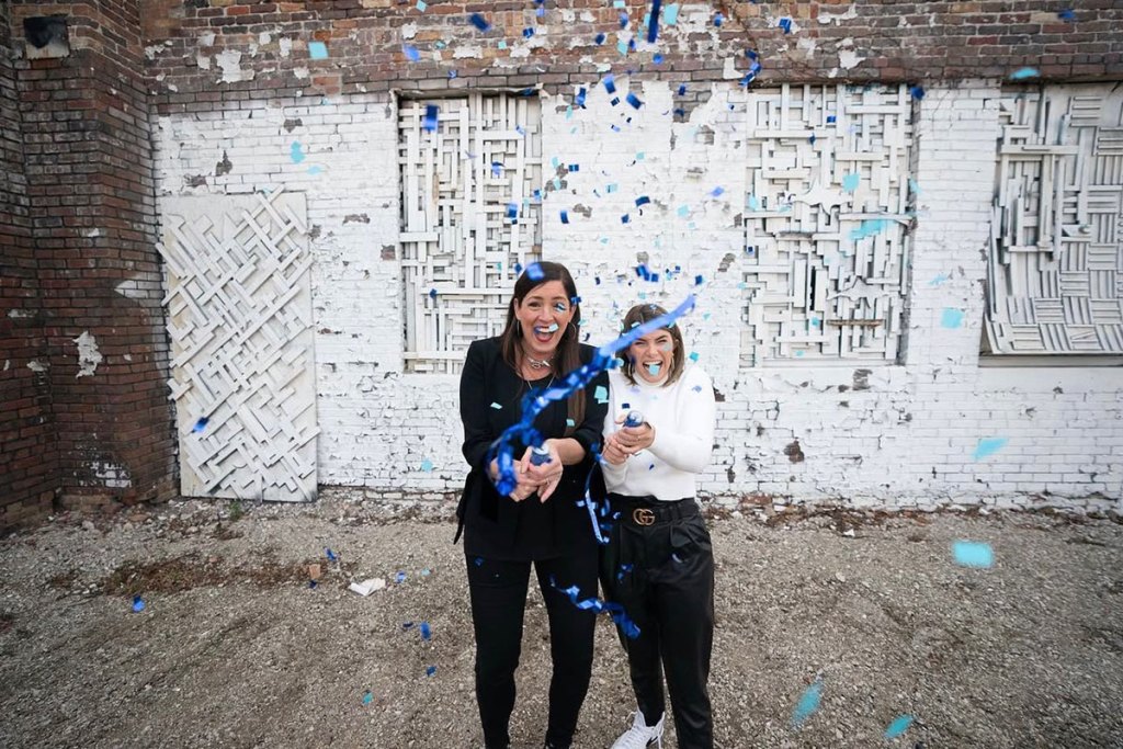 Two women stand outside and pop blue confetti towards the camera