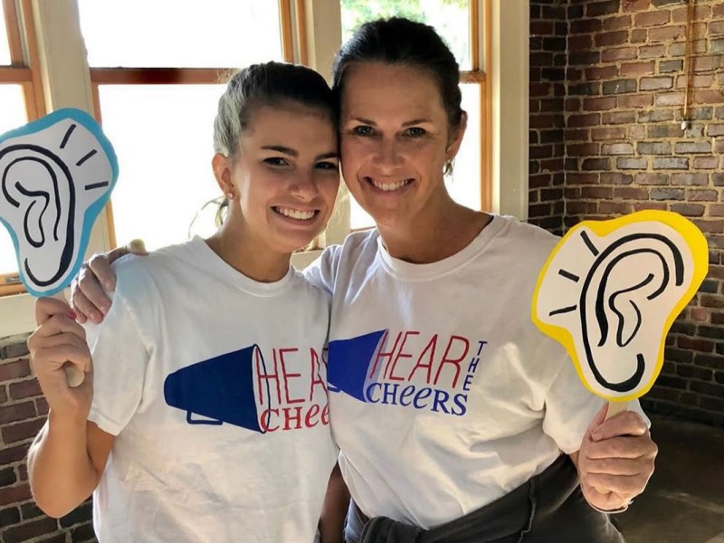 Two women wearing tshirts that read Hear the Cheers, holding up signs of ears