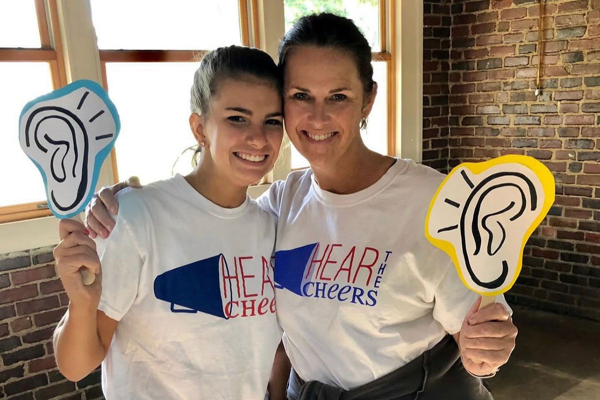Two women wearing tshirts that read Hear the Cheers, holding up signs of ears