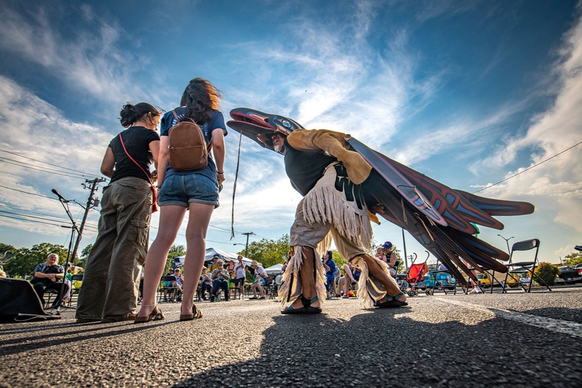 A man wearing a blue and red bird costume stands in the middle of people sitting around him