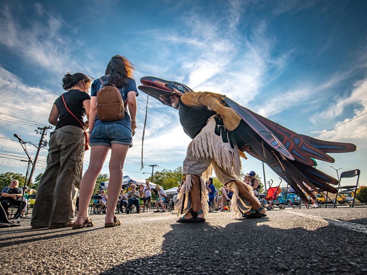 A man wearing a blue and red bird costume stands in the middle of people sitting around him