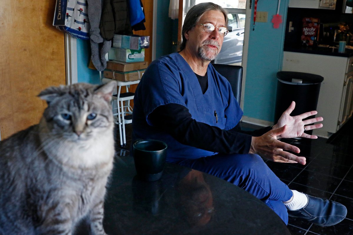 A man in blue scrubs sits in a kitchen and speaks. A grey and white cat sits in the foreground