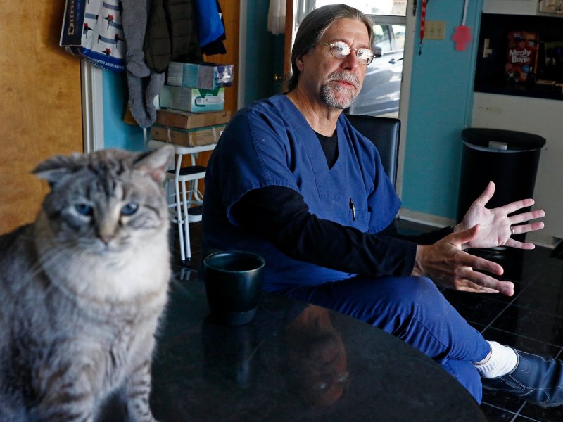 A man in blue scrubs sits in a kitchen and speaks. A grey and white cat sits in the foreground