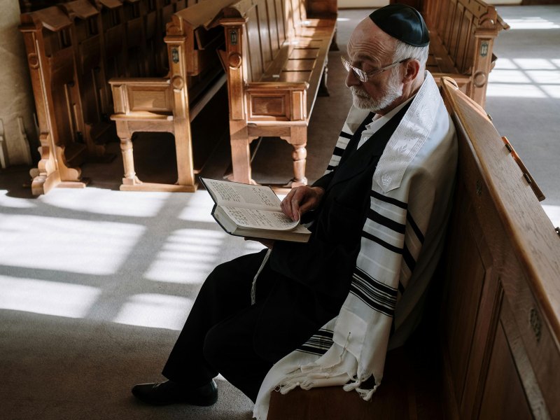An elderly man in Jewish garb sitting in a pew, open book in hand
