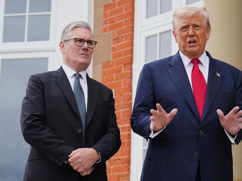 Keir Starmer stands on the left, listening, as Donald Trump speaks outside of a red brick building