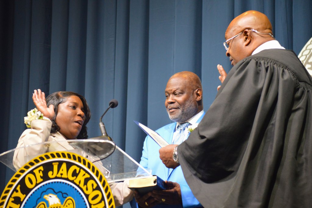 Lashia Brown wearing a tan suit stands with her pastor while being sworn in by a judge wearing a black robe