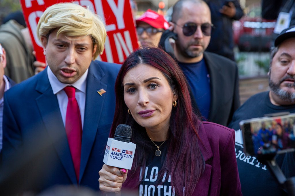 A woman holds a mic labeled Real America's Voice stands in a crowd, a person dressed like Donald Trump behind her