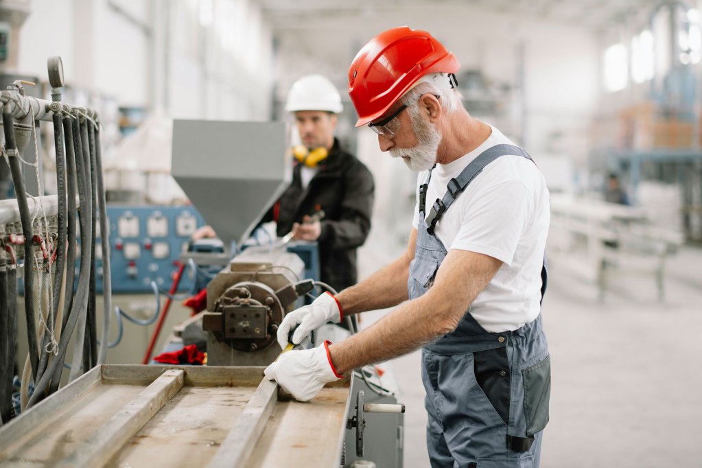 An older man uses a saw inside of a factory as another man works on another bench