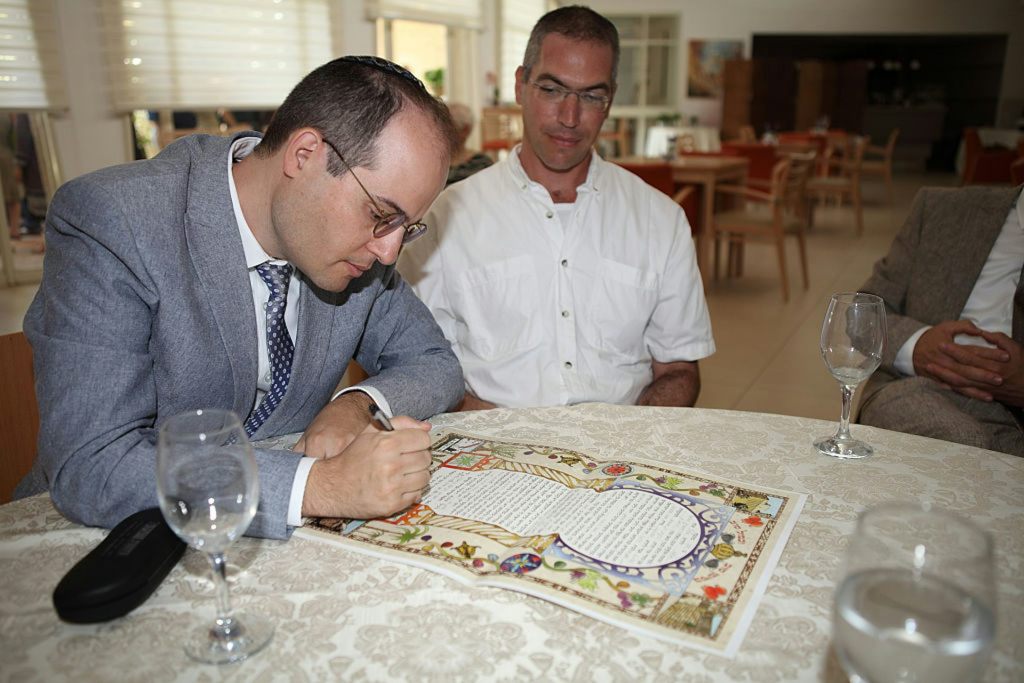A man in a blue-gray suit signs a colorfully decorated piece of paper as another man in a white shirt watches.