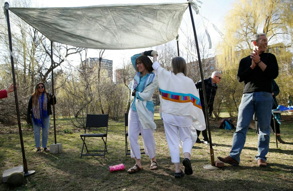 Two women wearing white clothes and prayer shawls dance under a simple canopy in a park as a few people look on