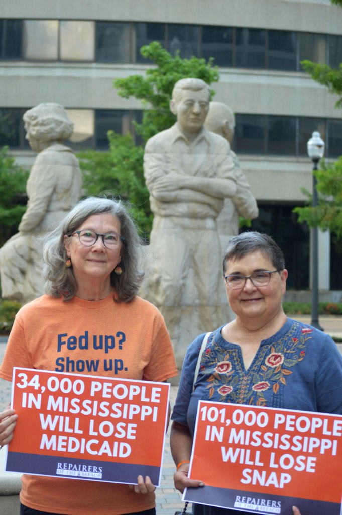 Two people stand outside before a statue holding signs that read '34,000 People in Mississippi Will lose Medicaid' and '101,000 People in Mississippi Will Lose SNAP'