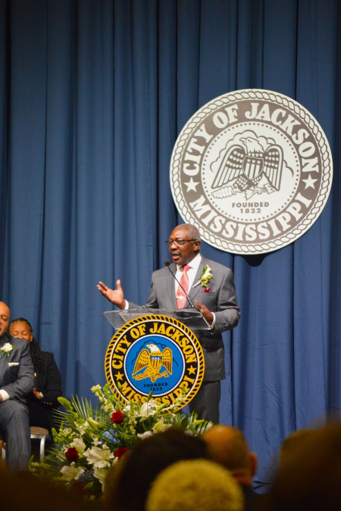 Mayor John Horhn wearing a grey suit with a red tie standing in front of a blue draped backdrop