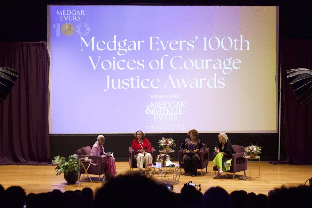 People seated on a stage, with a projected sign behind them that reads Medgar Evers' 100th Voices of Courage Justice Awards