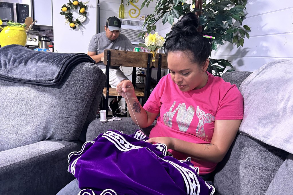 A woman in a pink top sits on her couch and sews on a purple and white garment
