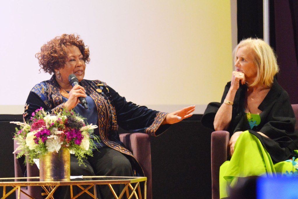 Reena Evers-Everette holding a mic wearing blue speaking to Kerry Kennedy wearing green on a stage