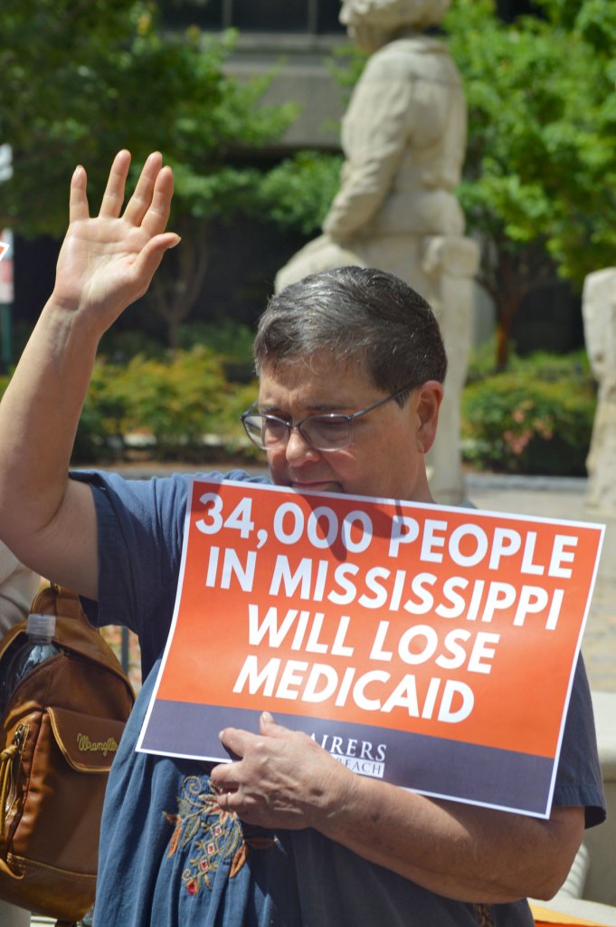 A person, one hand in air, holds a sign that reads '34,000 People in Mississippi Will lose Medicaid'
