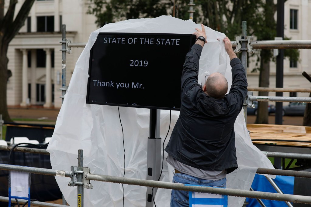 A man covers a teleprompter that reads 'State of the State 2019' with plastic sheeting in the rain