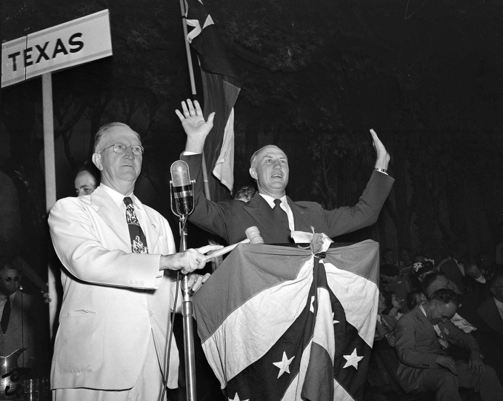 An old black and white photo of Strom Thurmond speaking at a US flag covered podium, arms up in the air to the crowd