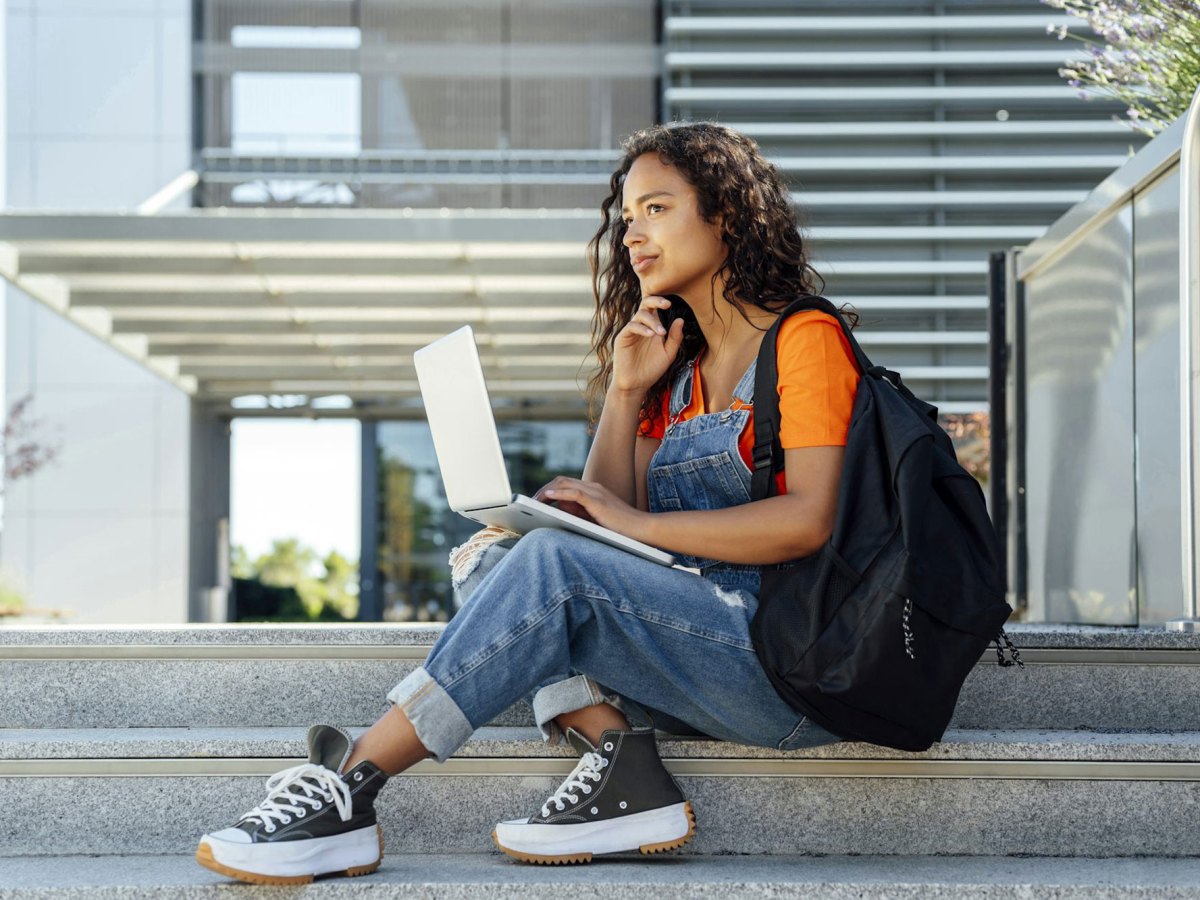 A student sitting on an exterior staircase by the school, laptop out