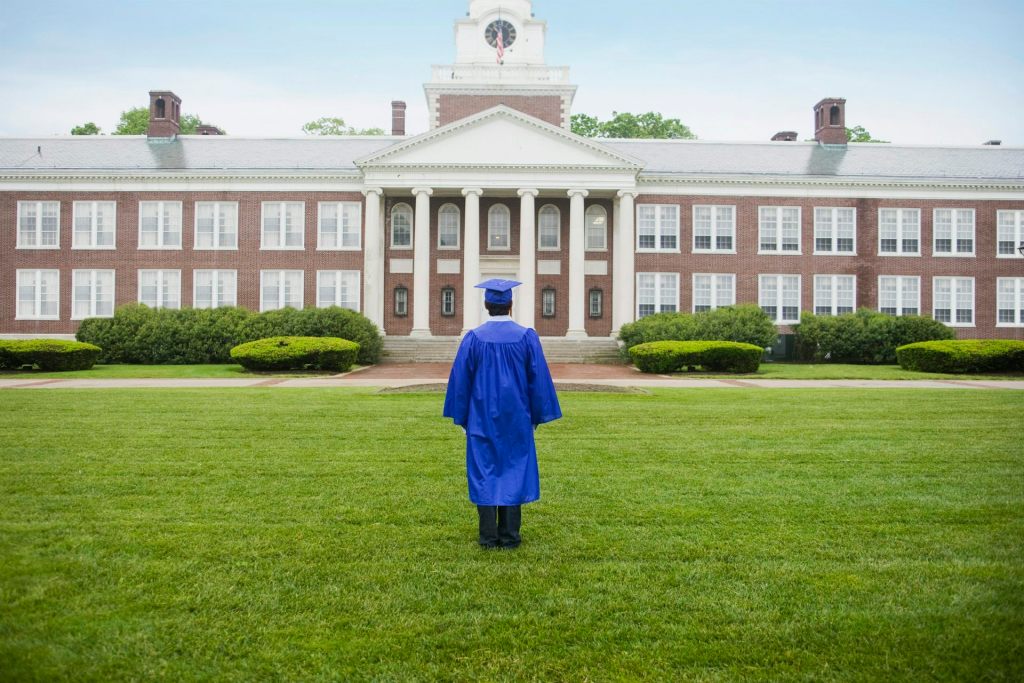 A student wearing a blue cap and gown stands on grass looking at a building."