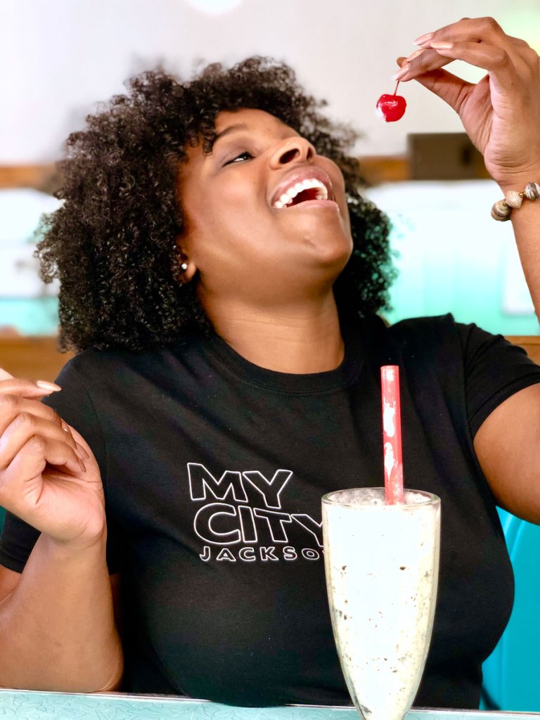 A woman in a black tshirt that reads My City Jackson holds a cherry aloft, ready to eat it, with a milkshake sitting in front of her