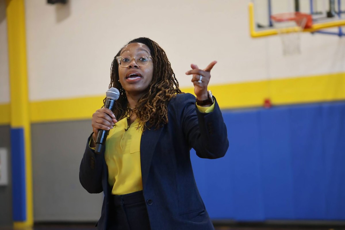 A woman in navy and yellow suit outfit speaks in a school gym, mic in one hand and pointing with the other