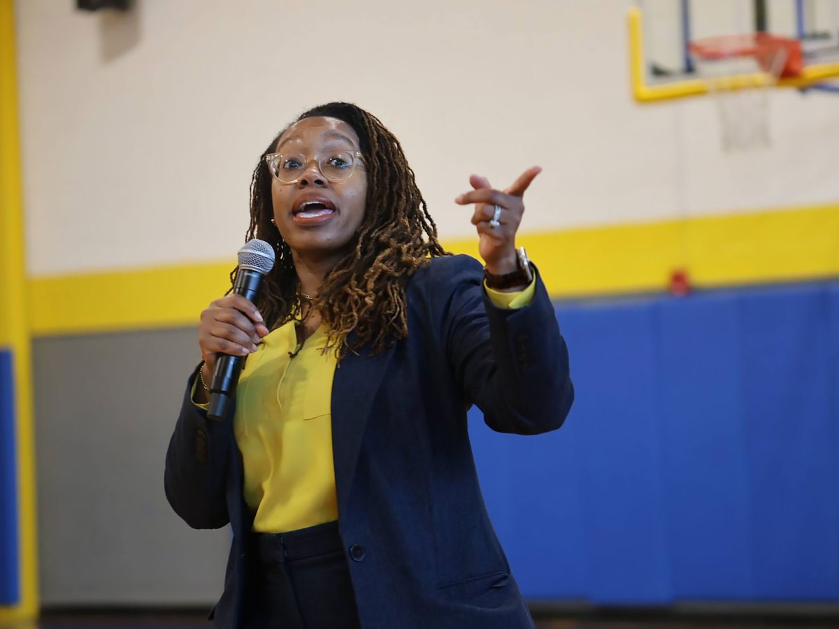 A woman in navy and yellow suit outfit speaks in a school gym, mic in one hand and pointing with the other