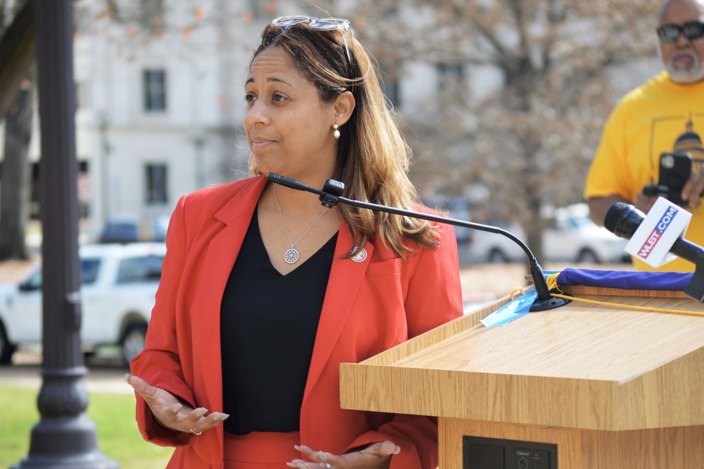 Zakiya Summers wearing a orange suit with a black shirt stands at a podium with a mic