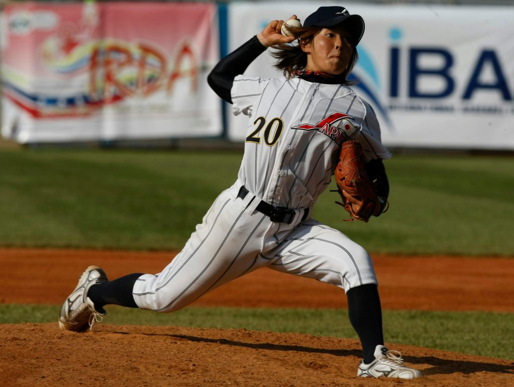 Young Japanese woman wearing a baseball uniform throws a pitch from a mound.