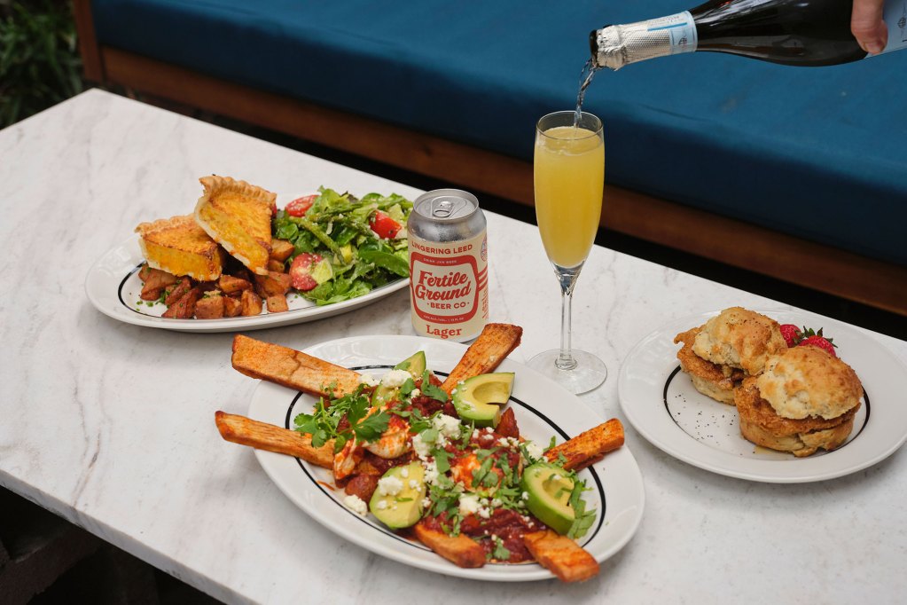 A hand is seen pouring champagne into a glass of mimosa. Three platters of food and a can of Fertile Ground beer sit around the table