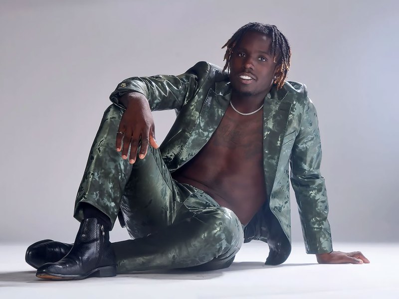 Singer Jamal Roberts sitting on the ground of a white backdrop wearing a green suit with no shirt