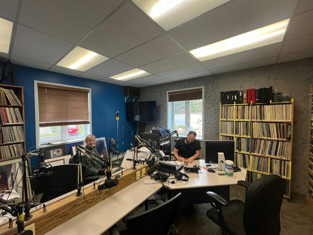 Two people inside a radio studio, sitting at a long table-desk combination