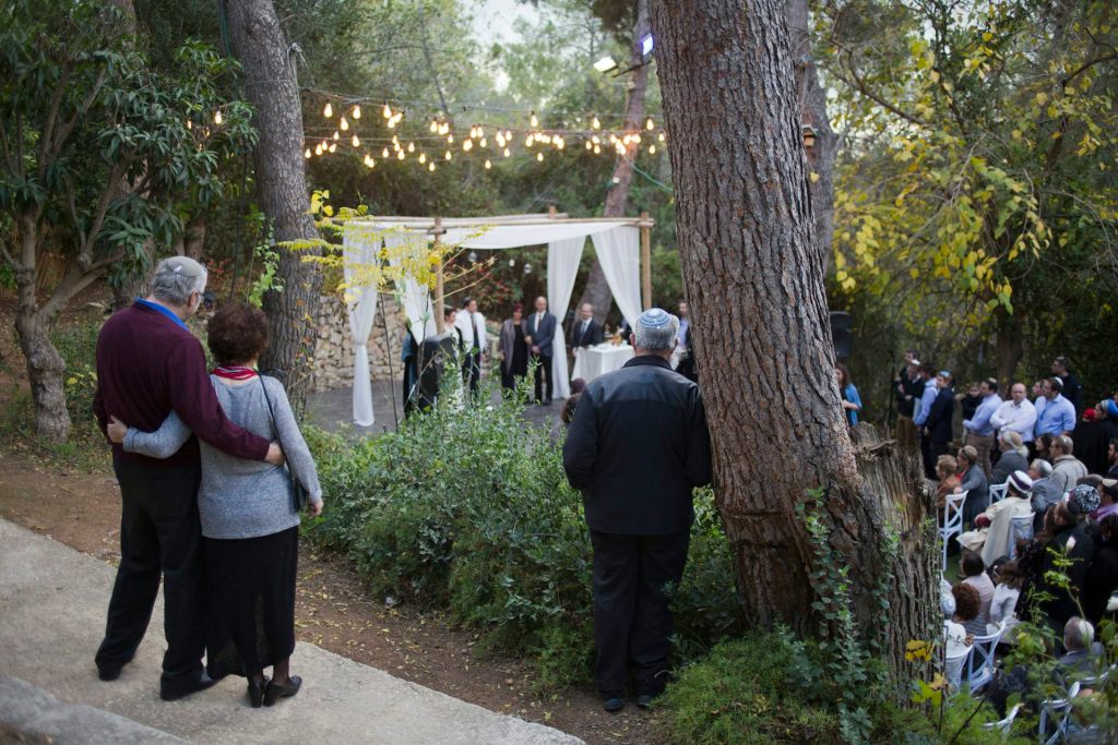 A couple embraces side by side as they observe a small wedding in a wooded area