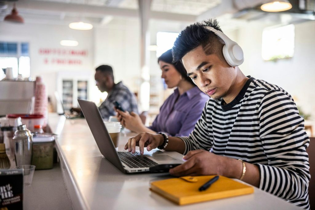 A man wearing headphones checks his phone while working on a laptop