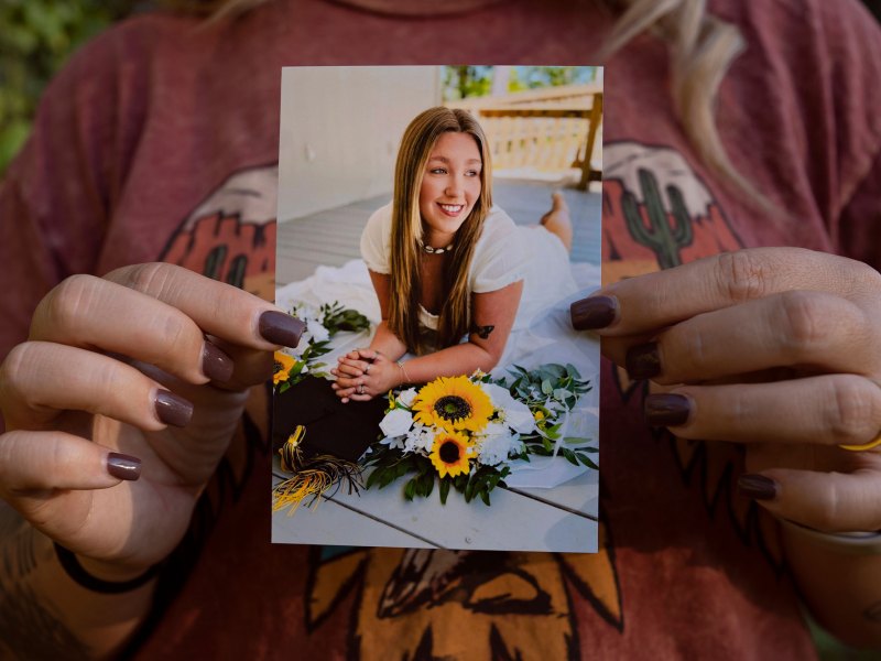 A pair of hands are seen holding a photo of a woman posed with sunflowers