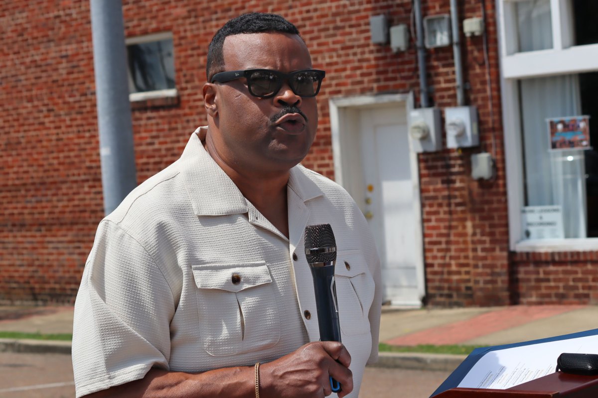 A man wearing sunglasses speaks on a microphone outside a red brick building