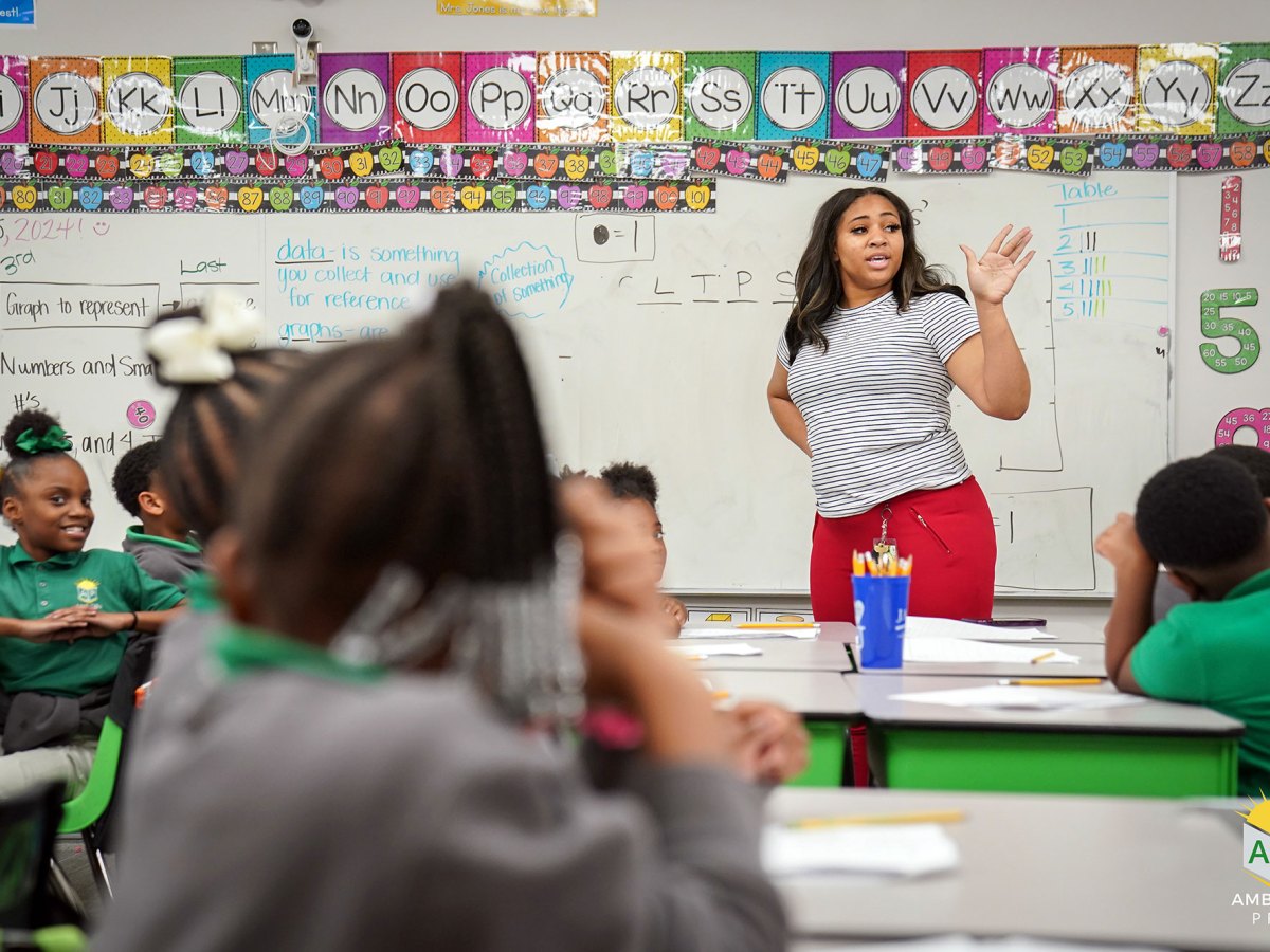 A teacher in a colorful grade school classroom teaches seated students all wearing school uniform shirts