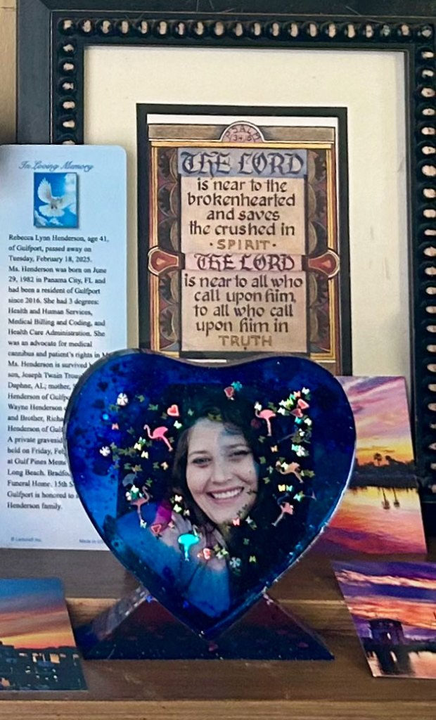 A picture of a woman in a blue heart-shaped frame sits in front of a framed canvas with a Bible verse and a copy of an obituary.