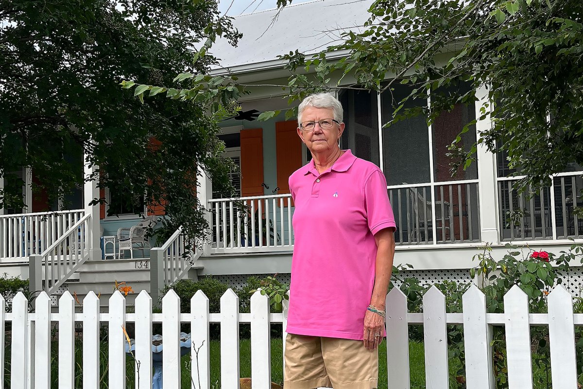 A woman in a pink shirt stands by the white fence of a home shaded by trees