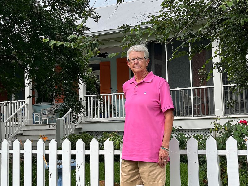 A woman in a pink shirt stands by the white fence of a home shaded by trees