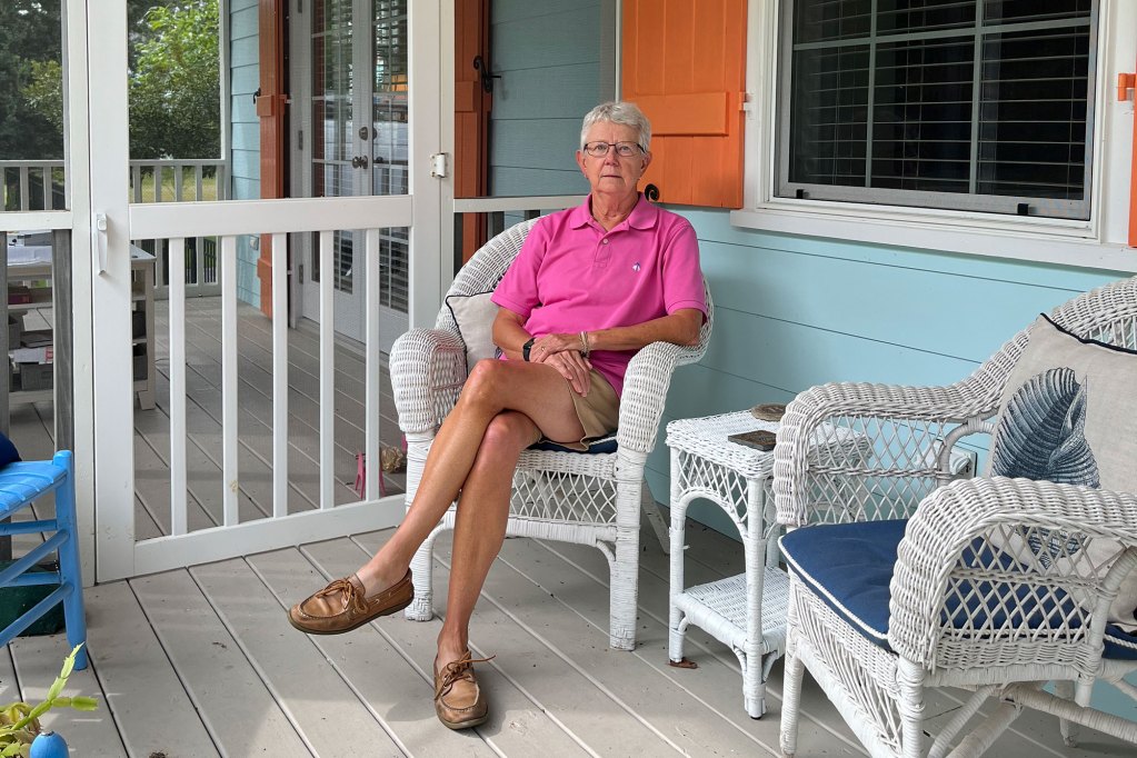 A woman in a pink shirt sits on the porch of a house with orange storm shutters