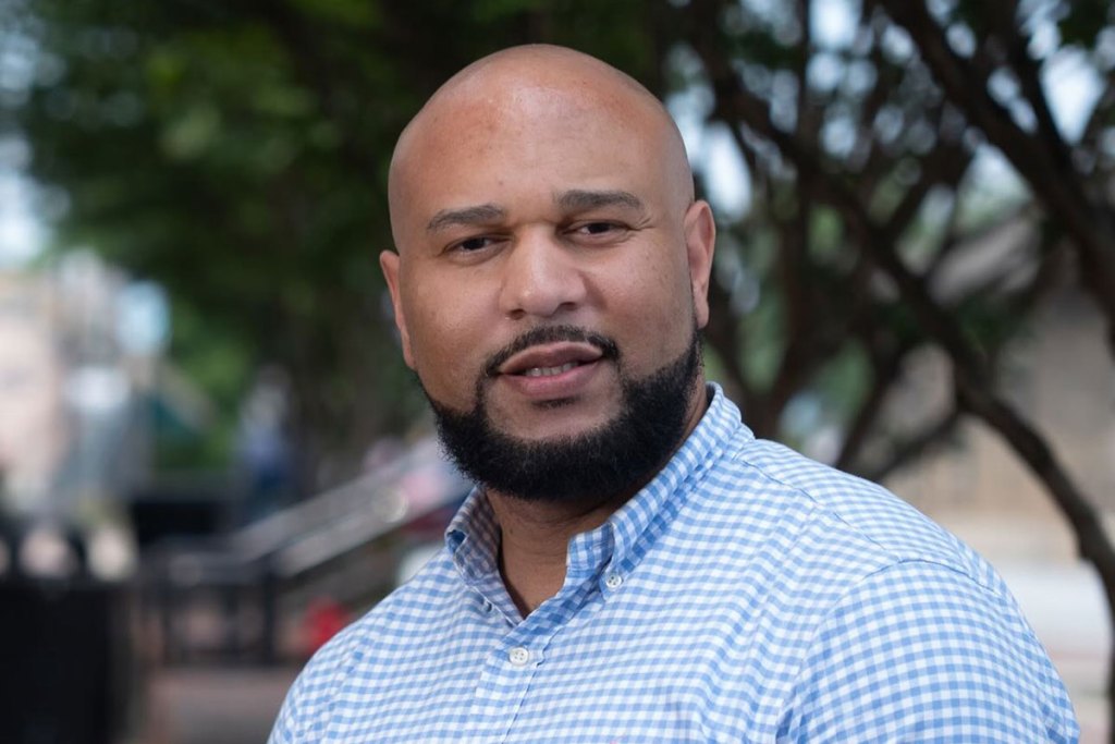 A bearded man in a white and blue checkered shirt standing outside