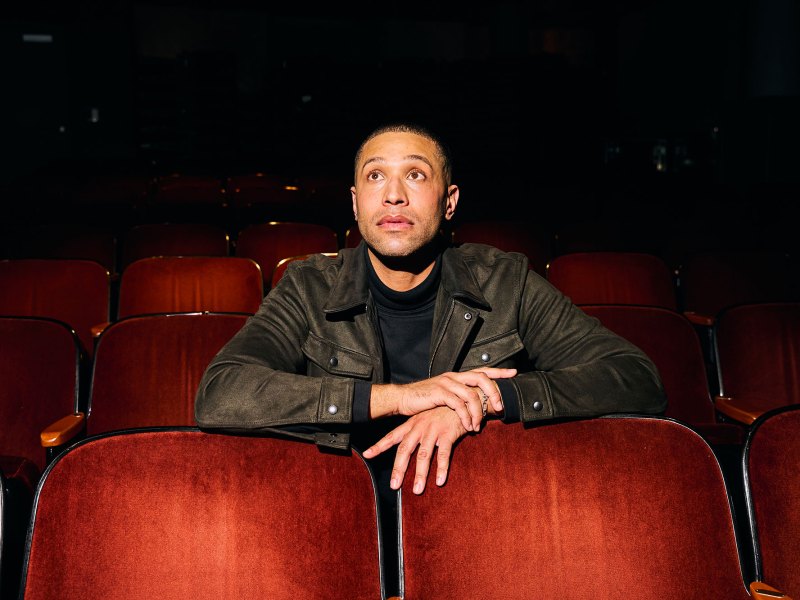 A man in a black jacket leans forward in a row of red velvet theater seats