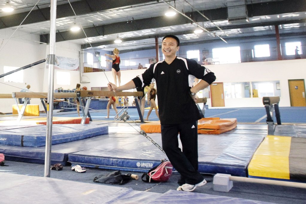A man in a black gym uniform leans against gym equipment as girls practice behind him