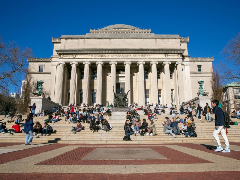 Students sit on the steps of a white columned building labeled The Library of Columbia University