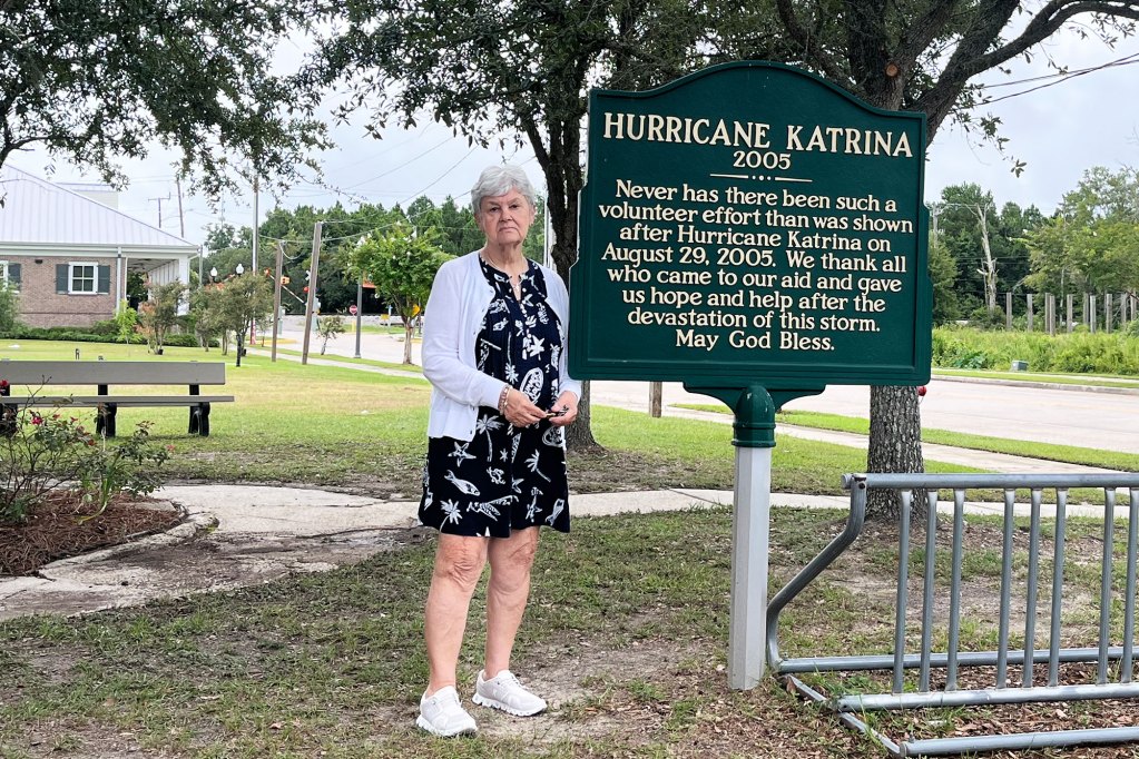 A woman stands in a park beside a sign 'Hurricane Katrina 2005. Never has there been such a volunteer effort than was shown after Hurricane Katrina on August 29, 2005. We thank all who came to our aid and gave us hope and help after the devastation of this storm. May God Bless.'