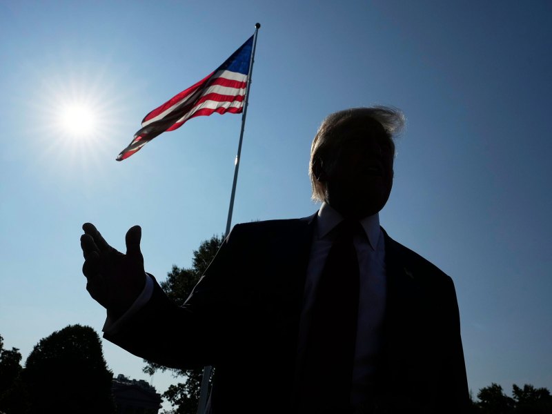 A dark silhouette of Donald Trump talking outside with the sun and a US Flag visible behind him