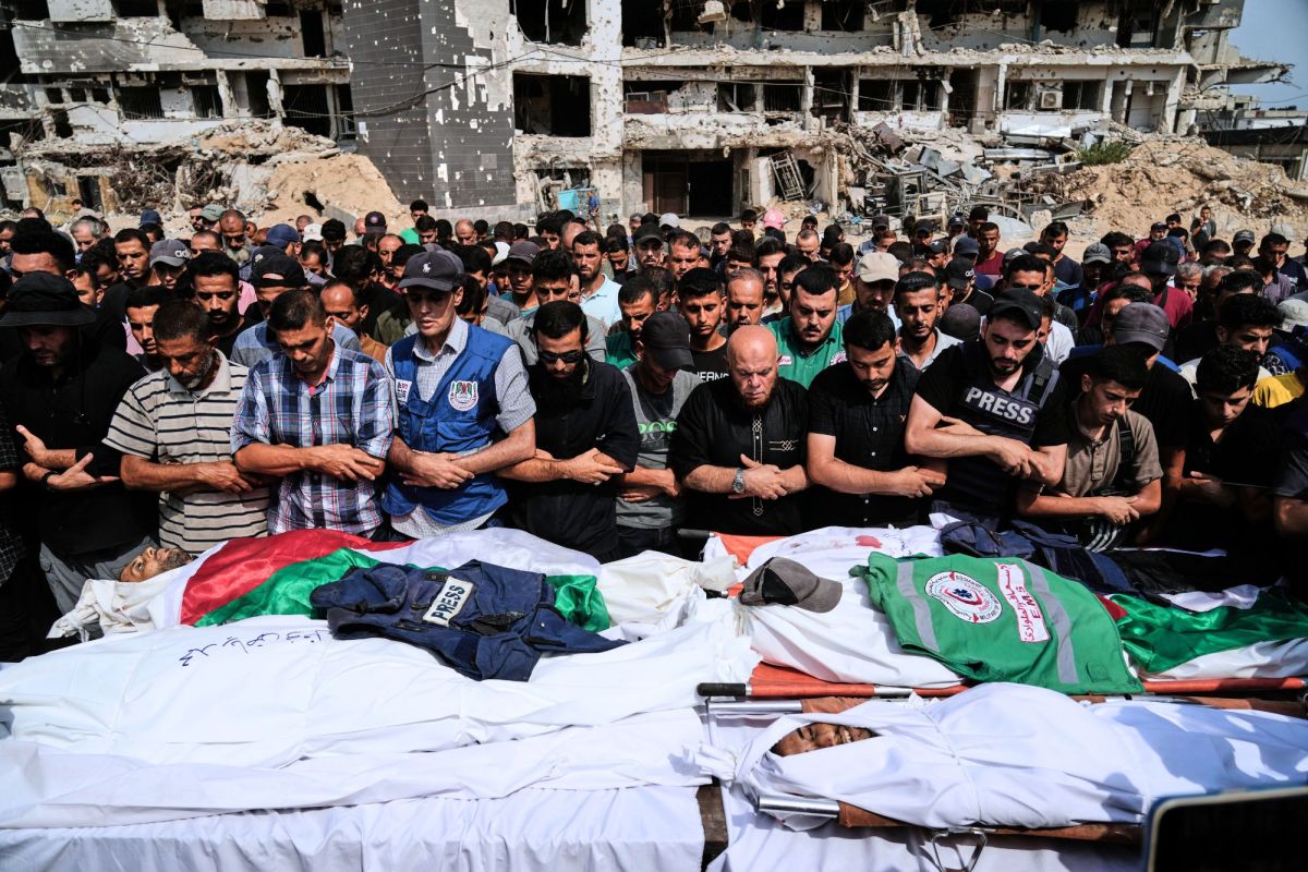 A large crowd prays over bodies covered in white sheets in a war-torn city