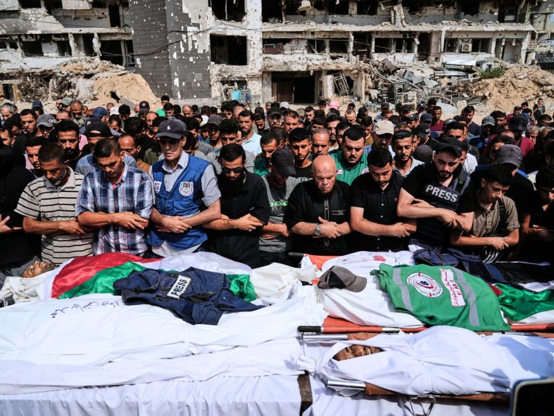 A large crowd prays over bodies covered in white sheets in a war-torn city